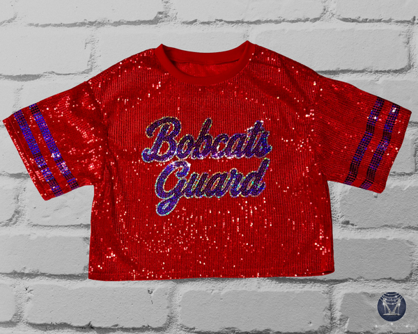 A red sequined short-sleeve jersey top shown from the front on a light gray brick background. The front reads "Bobcats Guard" in blue script in silver and blue bling with blue sleeve stripes.