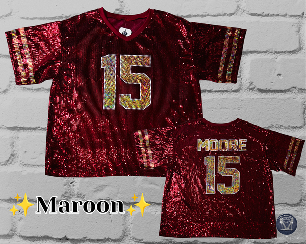 Two maroon sequined football jerseys displayed against a white brick wall background. The larger front view shows the number 15 in a glittery gold and silver pattern, and the smaller back view shows the name MOORE above the number 15.