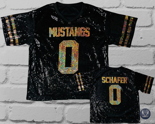 Two black short-sleeve football-style shirts with sequined lettering, shown against a white brick wall. The larger front view reads "MUSTANGS" above a large number 0, and the smaller back view reads "SCHAFER" above a number 0.