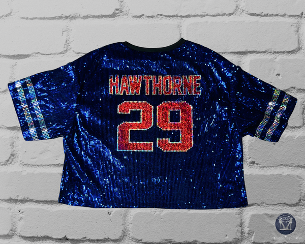 Back view of a blue sequined short-sleeve  on a light gray brick background. The back reads "HAWTHORNE 29" in red and silver block letters with silver sleeve stripes.