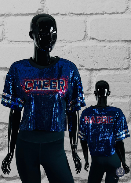 Two mannequins wearing blue sequin cheerleading outfits with 'CHEER' and 'MAGGIE' text against a white brick wall.