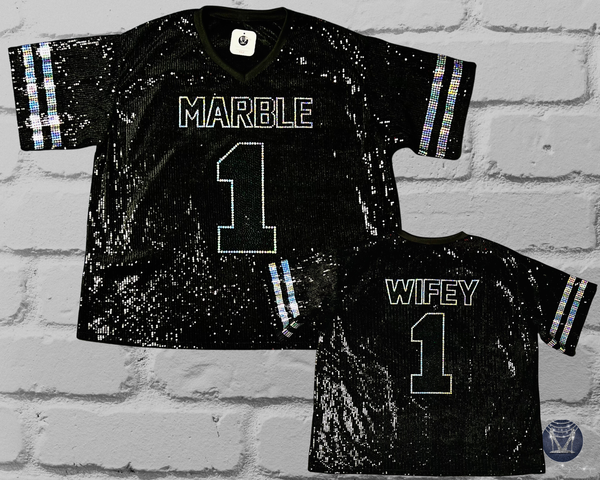 Two black sequined football jerseys displayed against a white brick wall background. The larger jersey shows the front with 'MARBLE' and the number '1' in white outlined lettering, while the smaller jersey shows the back with 'WIFEY' and the number '1'.