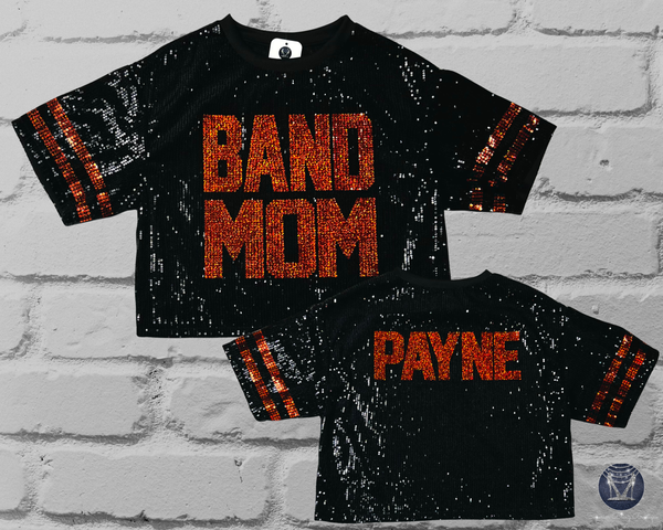 A black sequined short-sleeve mid crop top shown from both front and back against a white brick wall. The front reads 'BAND MOM' in large holographic orange letters, and the back reads ‘PAYNE' in similar orange lettering.