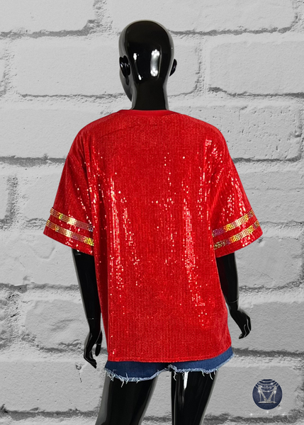 Red custom football-style sequin jersey shown from the back view with  striped bling gold sleeve accents, displayed on a black mannequin against a white brick wall.