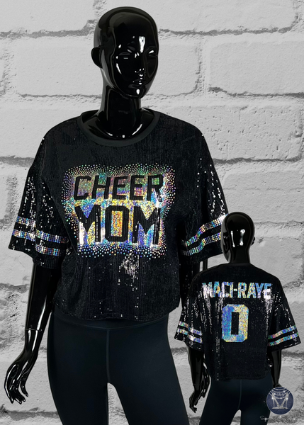 Black sequin 'CHEER MOM' shirt on a mannequin against a gray brick wall.